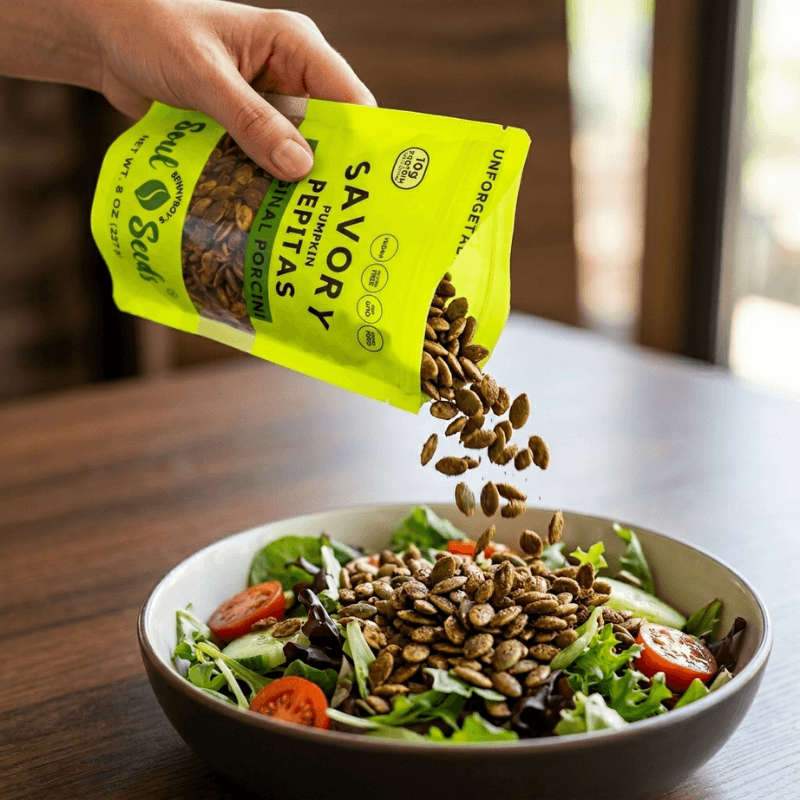 Hand pouring savory pumpkin seed pepitas from a yellow package into a salad.